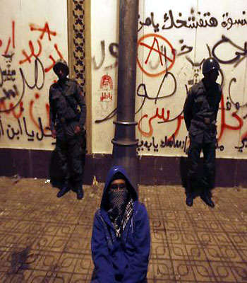 A masked anti-Mursi protester sits near Republican Guard soldiers standing guard in front of the presidential palace in Cairo. (R)