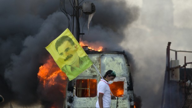 A protester holds a flag depicting jailed Kurdish rebel leader Abdullah Ocalan as a van burns during clashes at the Taksim Square in Istanbul Tuesday, June 11, 2013.Hundreds of riot police overran improvised barricades at Istanbul's Taksim Square on Tuesday, firing tear gas, rubber bullets and water cannons in running battles with protesters who have been occupying the area for more than a week. (AP Photo/Vadim Ghirda.)